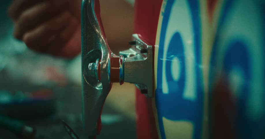 A person manually installs a silver metal truck onto a red skateboard deck by aligning the baseplate with mounting bolts over a blue and yellow graphic in a closeup shot