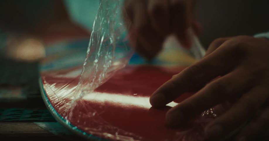 A close view of a person using a blade to cut and peel away protective plastic film from the surface of a colorful new skateboard deck on a workbench