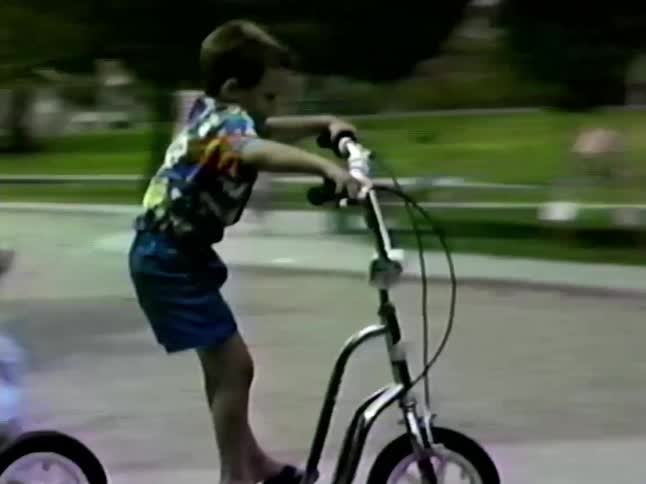 The video shows a young boy riding a large silver kick scooter with bicyclestyle wheels through a parkhe starts by riding across a paved area and transitions onto a grassy patch where he comes to a stop
