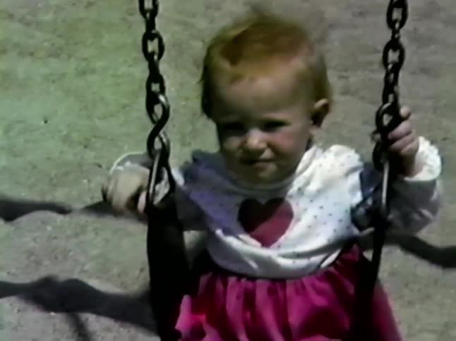 A young girl in a pink dress and a toddler in a white shirt with a red heart swing at a playgroundthe video is captured in a grainy vintage film style as the children smile while being pushed on the swings