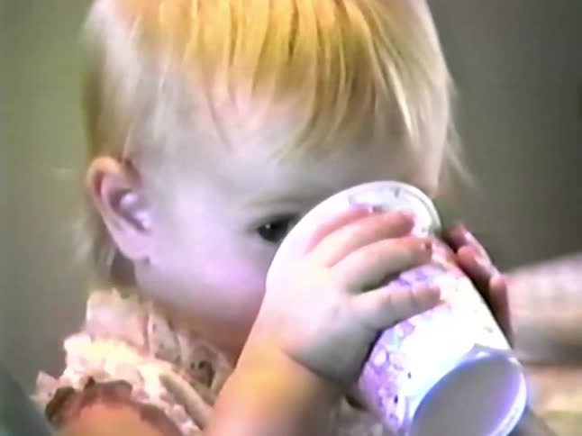 A toddler with blonde hair sits at a table and drinks from a small cup she then looks toward the camera with a wide excited expression as others gather around a pink cake