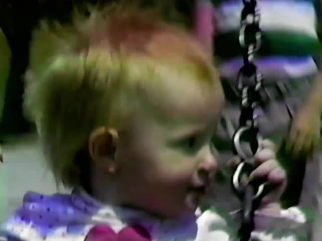 A boy in a blue striped shirt pushes a toddler girl in a heartpatterned outfit on a swing set at a sunny park