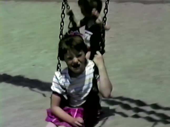 A young girl with a hair bow wears a striped shirt and pink skirt while smiling and swinging on a playground set during a sunny day while other children play on nearby swings