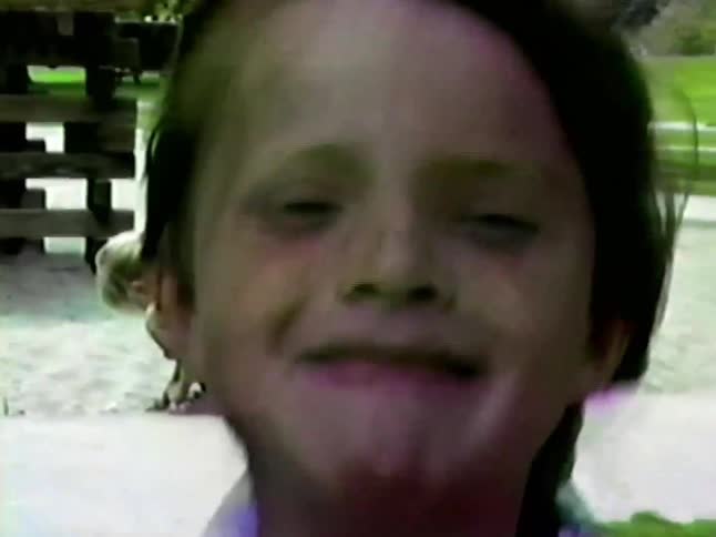 The video shows a young girl in a patterned buttondown shirt walking toward the camera at a playground behind her other children play near a sandbox and wooden climbing structure under the shade of green trees