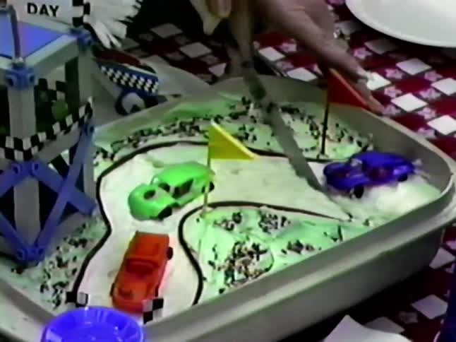 Adult woman cutting a cake decorated with toy cars with young girls smiling next to her and other children on a table decorated for a birthday party outdoors during day time