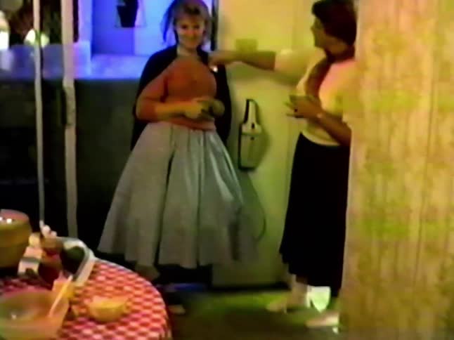Two women in 1950sstyle clothing stand in a kitchen one waves and holds a cup while the other shows off her full poodle skirt and veil as they laugh together while a boys head appears in front of the camera
