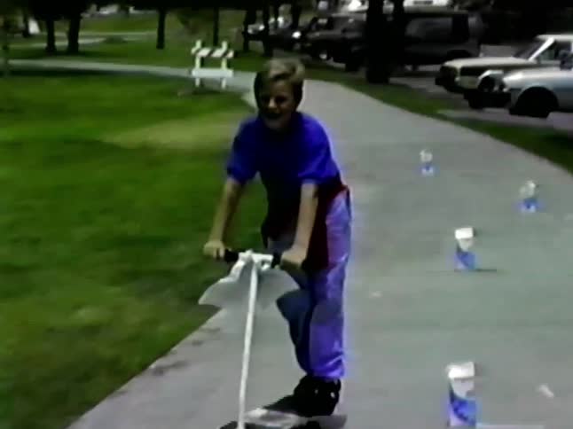 Boy almost falls while he races another on a scooter on a makeshift course in the park on a summer day