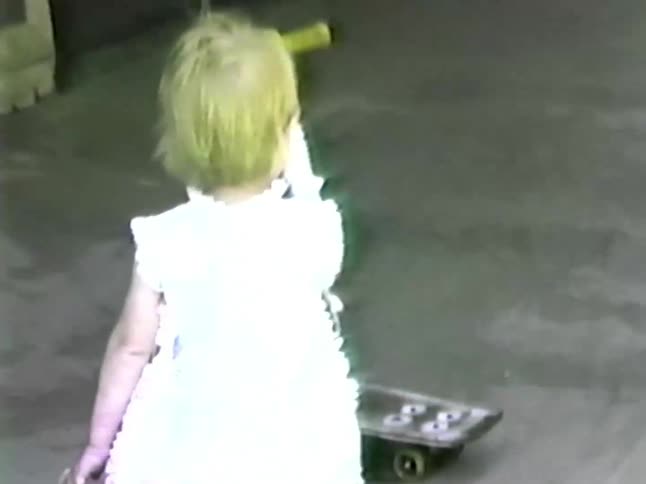 A toddler girl in a white dress sits on a small white and yellow scooter and laughs while playing in a concrete driveway