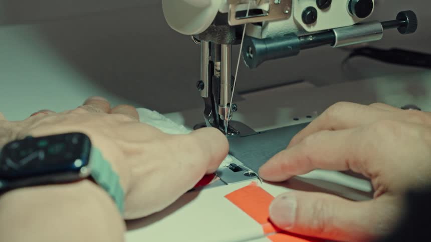 A close view captures the hands of a man using a sewing machine to stitch white fabric with orange tape