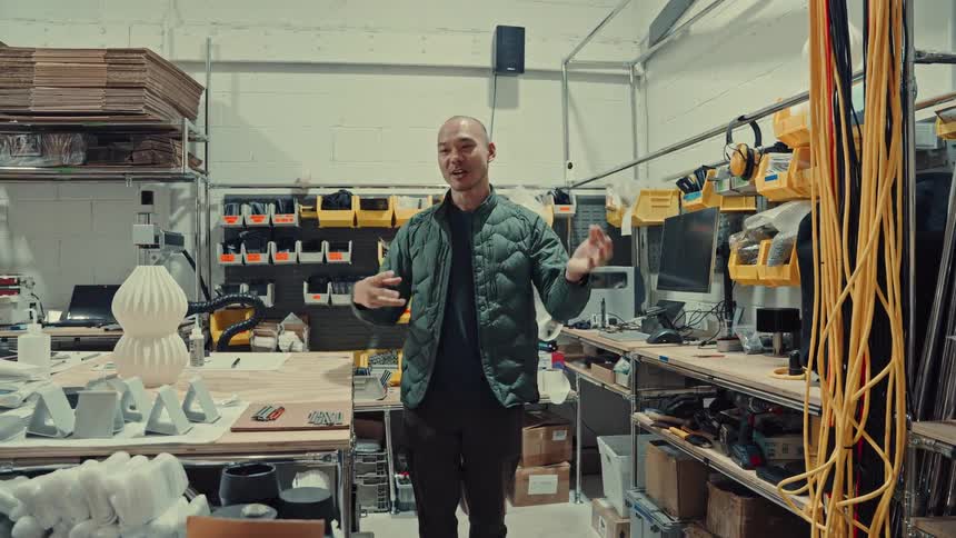 A man in a green jacket stands in a cluttered design workshop gesturing and explaining something while surrounded by tools shelving and industrial equipment