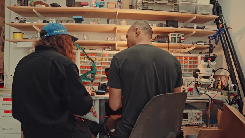 Two people working at an electronics bench with tools components organized in drawers and a microscope nearby
