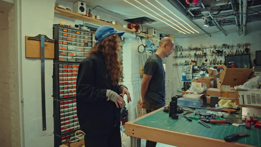 A woman with long curly hair and a blue cap talks and gestures with her hands while standing next to a man in a cluttered workshopthe workshop is filled with various tools storage bins and a large workbench covered in equipment and boxes
