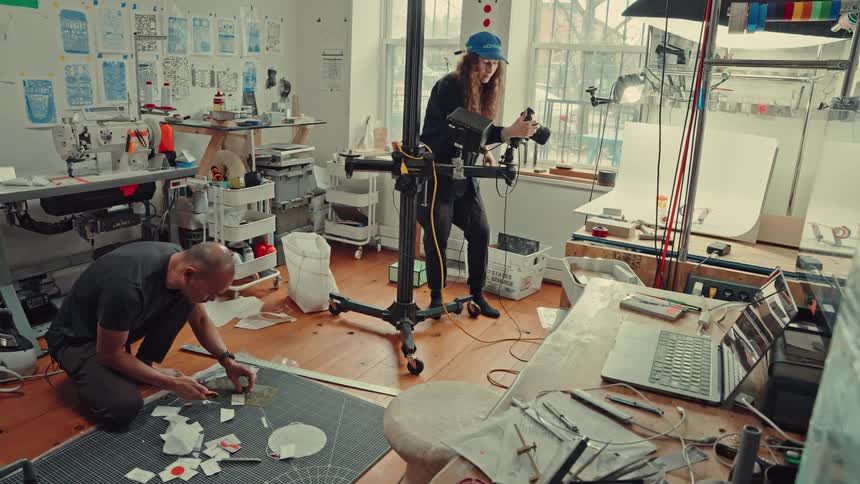 In a cluttered artists studio one person kneels to assemble a craft on the floor while another films the process with a large camera on a rolling stand