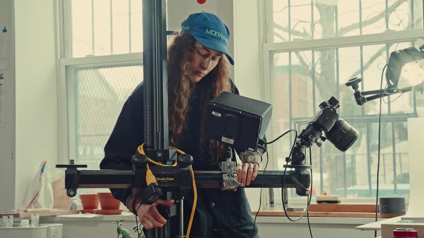 A woman with long curly hair wearing a blue cap adjusts a camera mounted on a large professional studio standthe scene takes place in a brightly lit room with a large window in the background showing a wintery outdoor view
