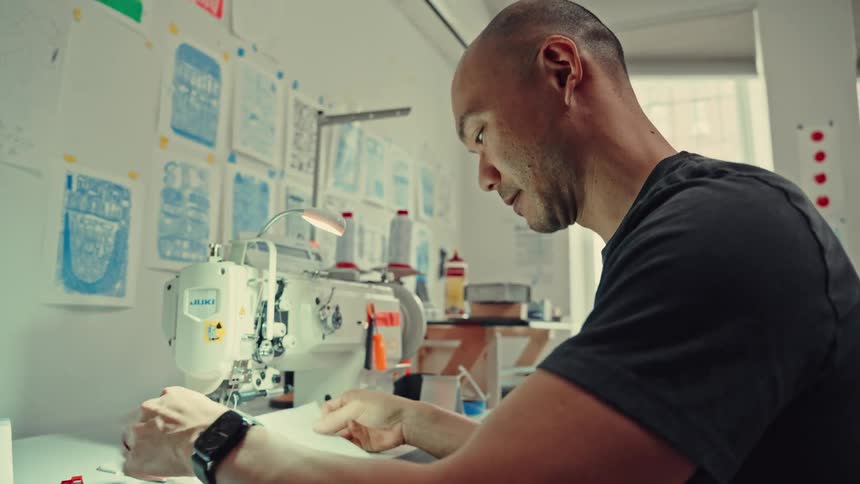 A man reaches into a white bag to retrieve fabric before sitting at an industrial sewing machine to carefully align and stitch the material in his workshop which is decorated with technical drawings and sketches on the wall