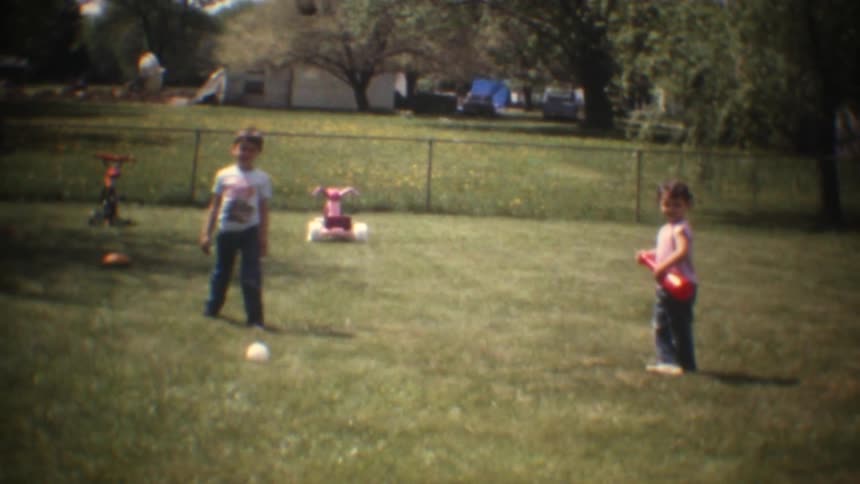 Young male child throwing a ball and young female toddler trying to hit it with a bat and missing on the lawn of a trailer park outdoors during day time