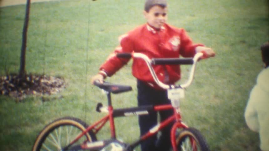 In this vintage video two children ride their bicycles along a residential driveway while a young girl waves happily at the camera as they pedal an adult carrying papers briefly walks across the foreground momentarily obscuring the view of the childre