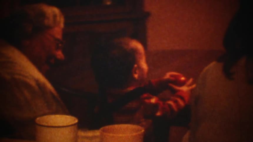 Elderly woman with a baby on his lap playing with an apple by rolling it over a table and smiling next to other people sitting on the table and smiling as well indoors during night time