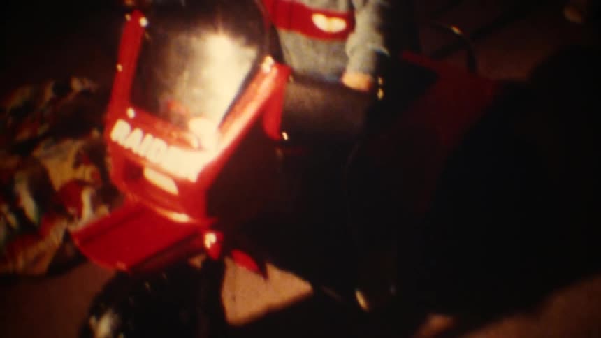 A young boy in a sweater sits on a red toy motorcycle and waves his hand in a room with vintage film grain