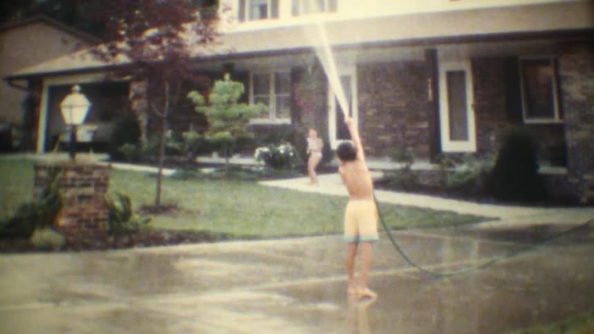 Two kids play with a water hose on the driveway on a sunny day the boy changing the water flow and sending the girl running away laughing as the spray chases her
