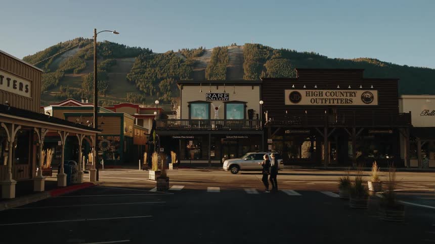 Downtown area in a rural town with people and cars passing by and a hill covered in trees on the background outdoors during sunset time