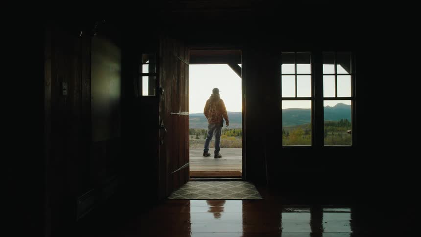 Young adult man walking through an open door and standing on the porch looking around at a natural landscape on a rural area indoors during day time