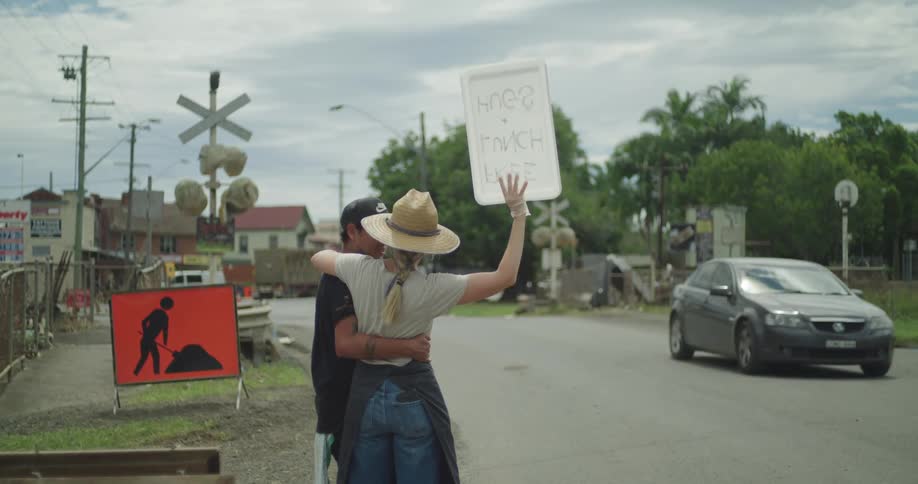 A couple stands on the side of the road at midday hugging as the woman holds a sign while a sedan drives past them