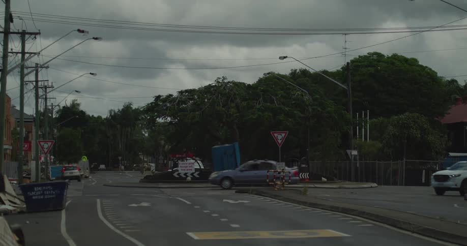 Various cars and a pickup truck navigate a suburban road intersection and roundabout under a heavy overcast sky while road signs and utility poles line the street surrounded by lush green trees