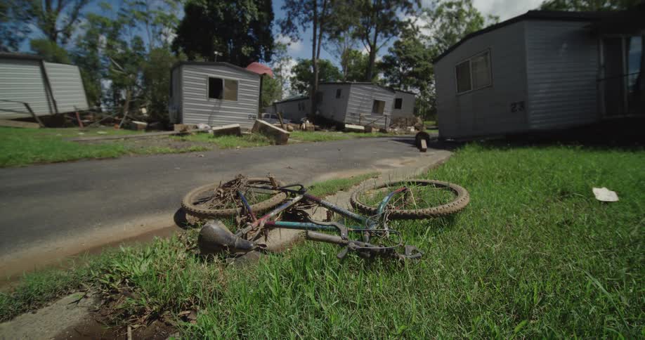 A bicycle lies on the side of a sunny street with buildings lining both sides of the road as bright daylight casts clear shadows across the pavement