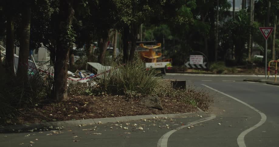 A white van and a yellow utility truck drive past a large pile of discarded furniture and household waste sitting on a landscaped curb near a busy urban intersection during the day