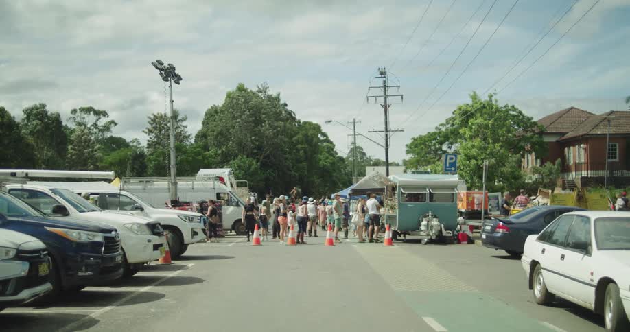A crowd of people gathers in a parking lot between lines of parked cars and a small blue food trailerbright daylight illuminates the outdoor scene which features orange traffic cones and tall light poles in the background