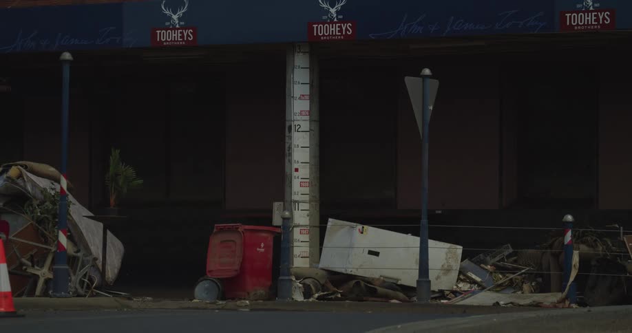 Cars drive past a building with a flood level gauge on the wall and a large pile of debris including a white appliance and red bin on the sidewalk