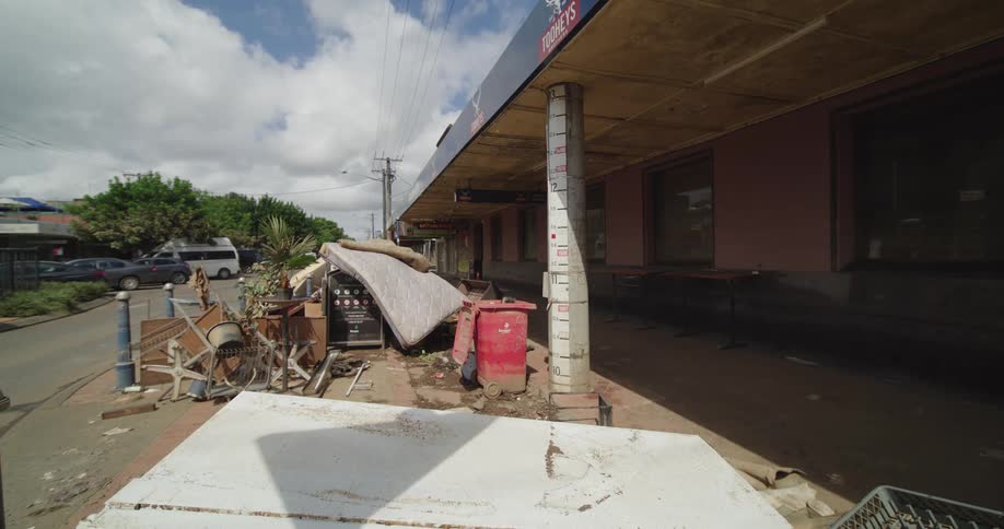 A sidewalk is covered in debris and discarded furniture outside a shop with cars passing by on the street under a cloudy sky with trees in the back