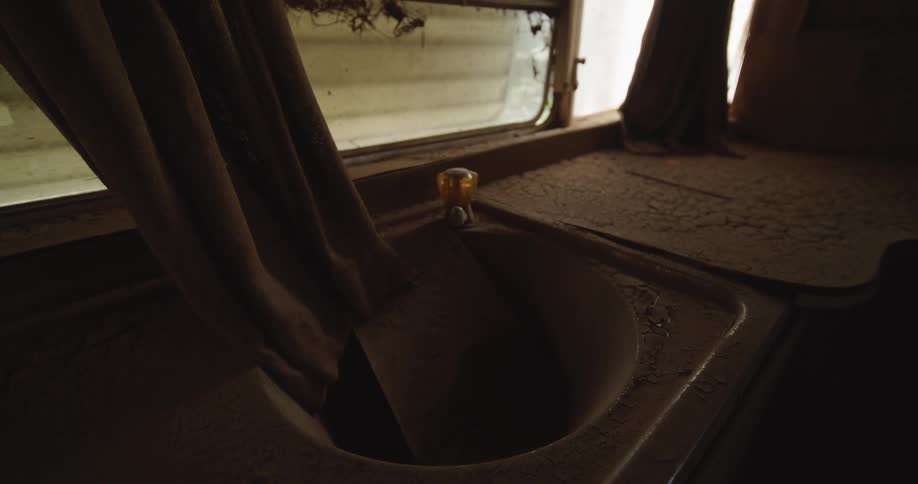 Close view of a sink covered in thick cracked dust inside an abandoned trailer with dim light entering through a curtained window