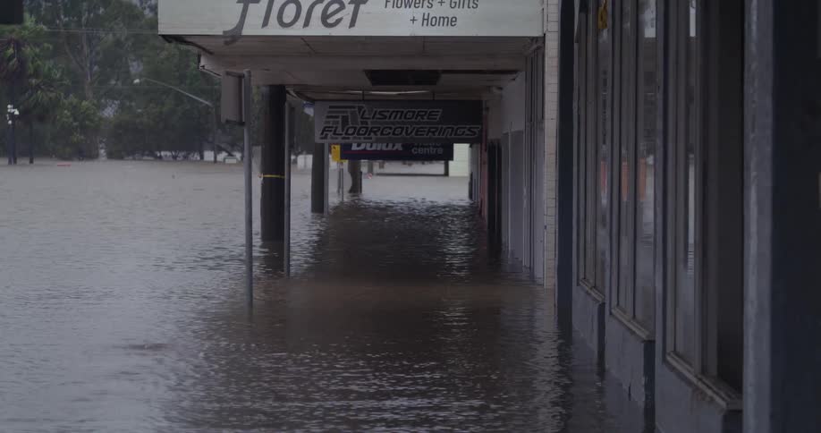 Murky floodwaters have completely submerged a street and the sidewalk in front of several storefrontssignage for businesses like floret and lismore floorcoverings is visible above the rising water line