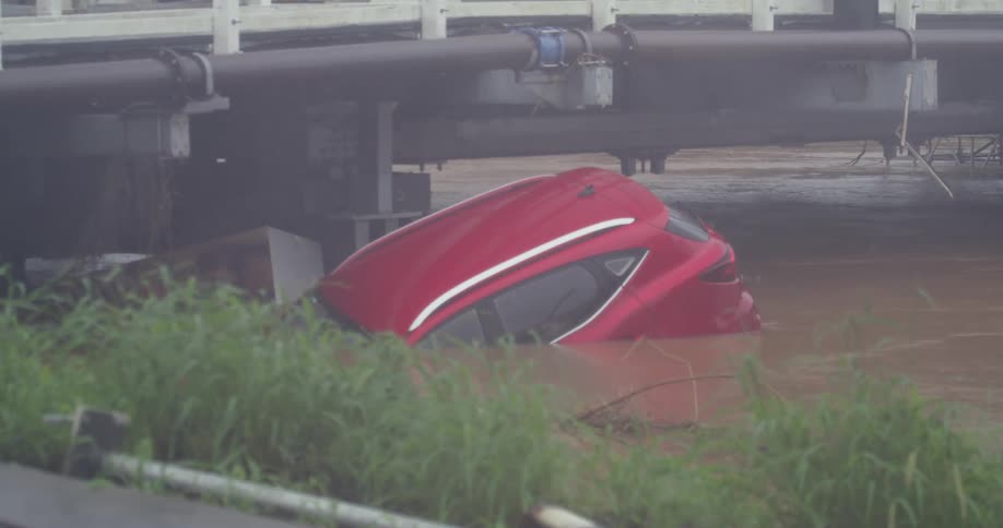 Red car partially submerged in muddy water beneath a bridge structure