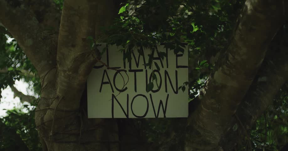 A white sign reading climate action now is posted on a large gnarled tree before the camera pans quickly away to reveal police officers and people gathered in a park setting