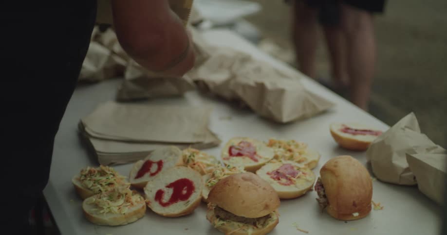 Men with tattoos work at a white table outdoors assembling burgers with coleslaw and sauce before placing them into brown paper bags for a community meal service