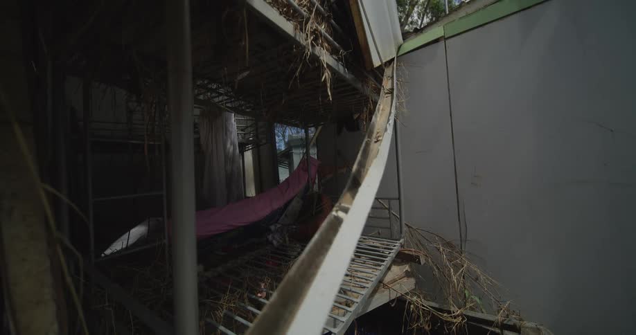 Inside a damaged partially collapsed structure a narrow metal walkway cluttered with debris and dried vegetation leads past torn panels and exposed framing toward a dim interior