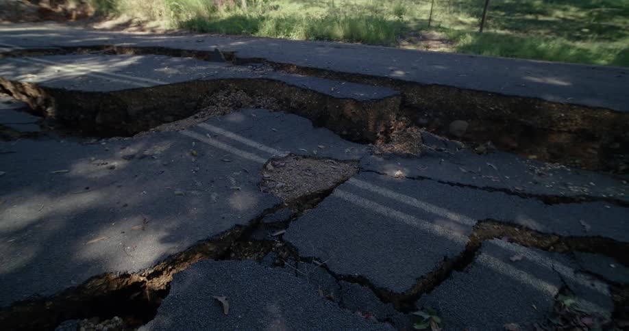A paved road is severely fractured and buckled with large chunks of asphalt displaced and sinking into deep fissures the damage reveals the underlying earth and gravel beneath the broken surface in a wooded area