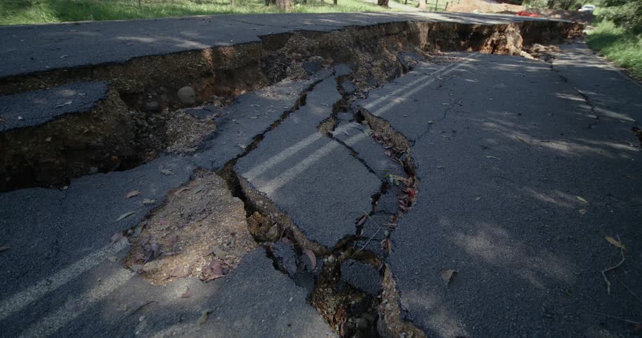 A tarred road severely damaged by an earthquake or landslide with wide cracks and a dramatic drop in elevation all illuminated by the bright midday sun
