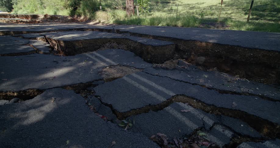 A rural asphalt road lies violently fractured and sunken into jagged sections with deep cracks and displaced slabs cutting across faded lane markings