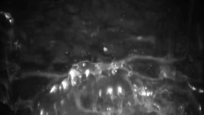 In a black and white closeup shot a viscous liquid churns and boils rapidly creating a dense layer of translucent bubbles that stretch and burst while shimmering under a bright light source against a dark background