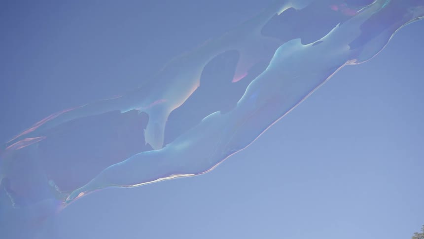 A closeup view shows large iridescent soap bubbles floating and stretching across a clear blue sky before they eventually drift out of frame