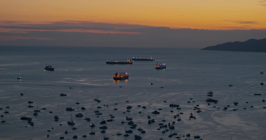 Large cargo ships and many small boats float in a harbor during a golden sunset with their lights glowing against the dark water and a mountain silhouette visible in the background under an orange and purple sky