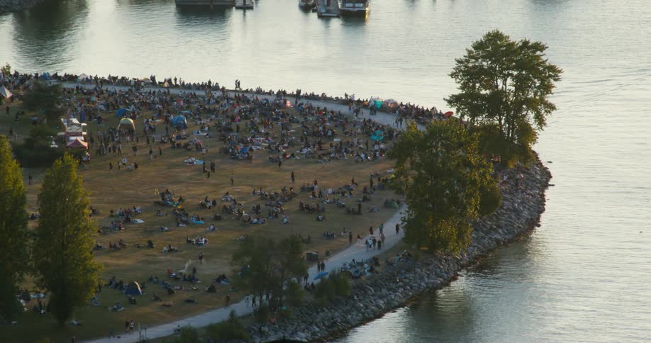Crowd gathered on grassy peninsula surrounded by water during daytime event with tents and boats