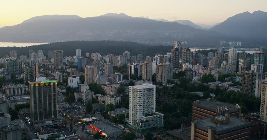 The video shows an aerial view of a dense cityscape nestled between a body of water and a mountain range during twilightwarm golden light from the setting sun illuminates the apartment buildings and the forestcovered park in the distance