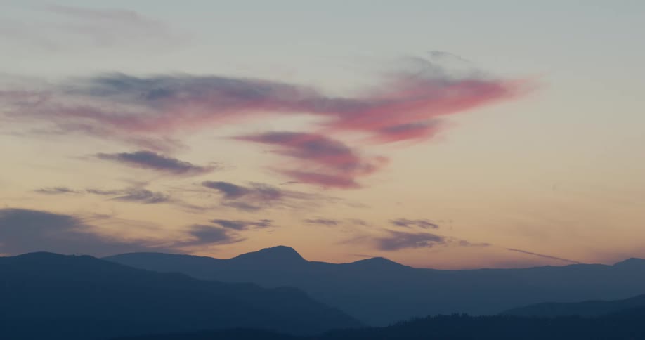 Soft pink and orange clouds drift slowly across a darkening twilight sky above the jagged blue silhouettes of a distant mountain range