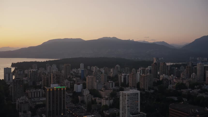 An aerial view shows a dense city skyline at dusk with numerous apartment buildings and skyscrapers stretching toward a body of waterin the background the silhouettes of large mountains are visible under a soft orange and purple sunset sky