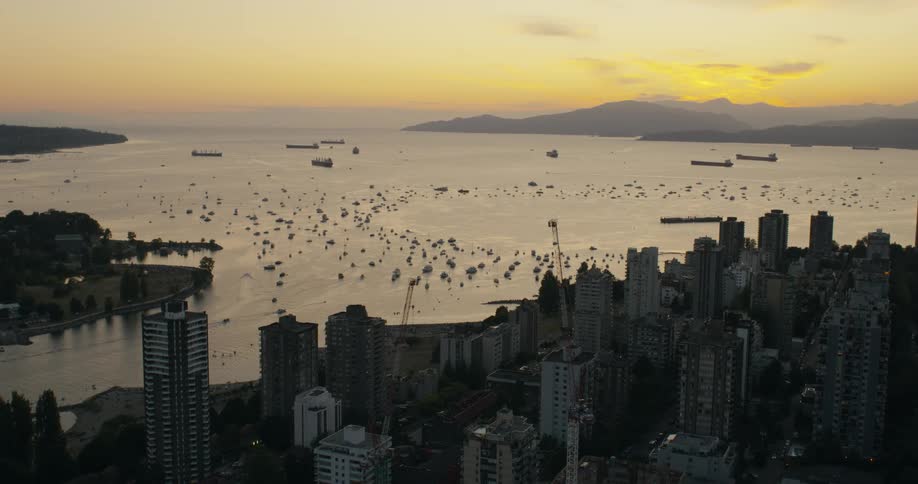 An aerial view shows a dense city skyline overlooking a calm bay filled with numerous small boats and large tankers under a golden sunset sky that silhouettes distant mountains and local highrise architecture